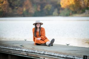 Person in orange sweater sitting on wooden dock by lake surrounded by fall foliage.