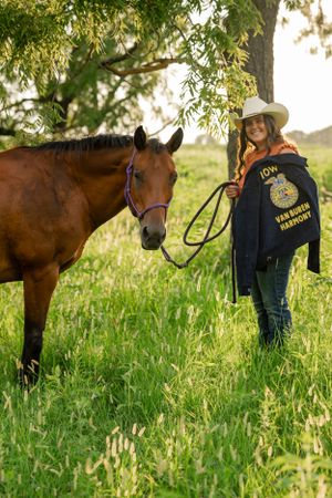 FFA member in official jacket standing with brown horse in grassy field.