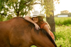 Person in cowboy hat hugging brown horse in sunlit field.