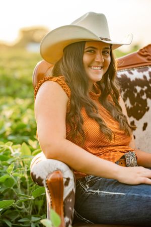 Person in cowboy hat and orange top sitting with spotted cowhide in field at sunset.
