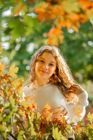 A person in a cream sweater smiles among fall foliage with orange leaves and bokeh lighting.