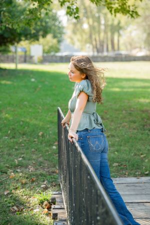 Casual pose leaning against black railing on wooden bridge in park setting.