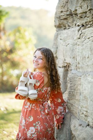 A floral dress-clad figure holds baby shoes while standing against a rustic stone wall in warm sunlight.