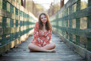 Someone in a red floral dress sits cross-legged on a wooden bridge with railings.