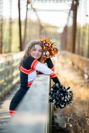 A cheerleader in uniform poses with pom poms on a rustic bridge during golden hour.