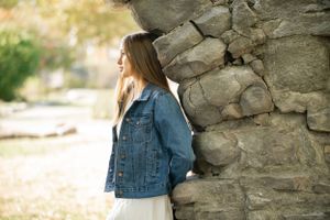 Figure leaning against weathered stone wall wearing denim jacket in sunlit outdoor setting.
