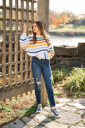 Figure wearing colorful striped sweater and jeans poses by wooden fence in autumn garden.