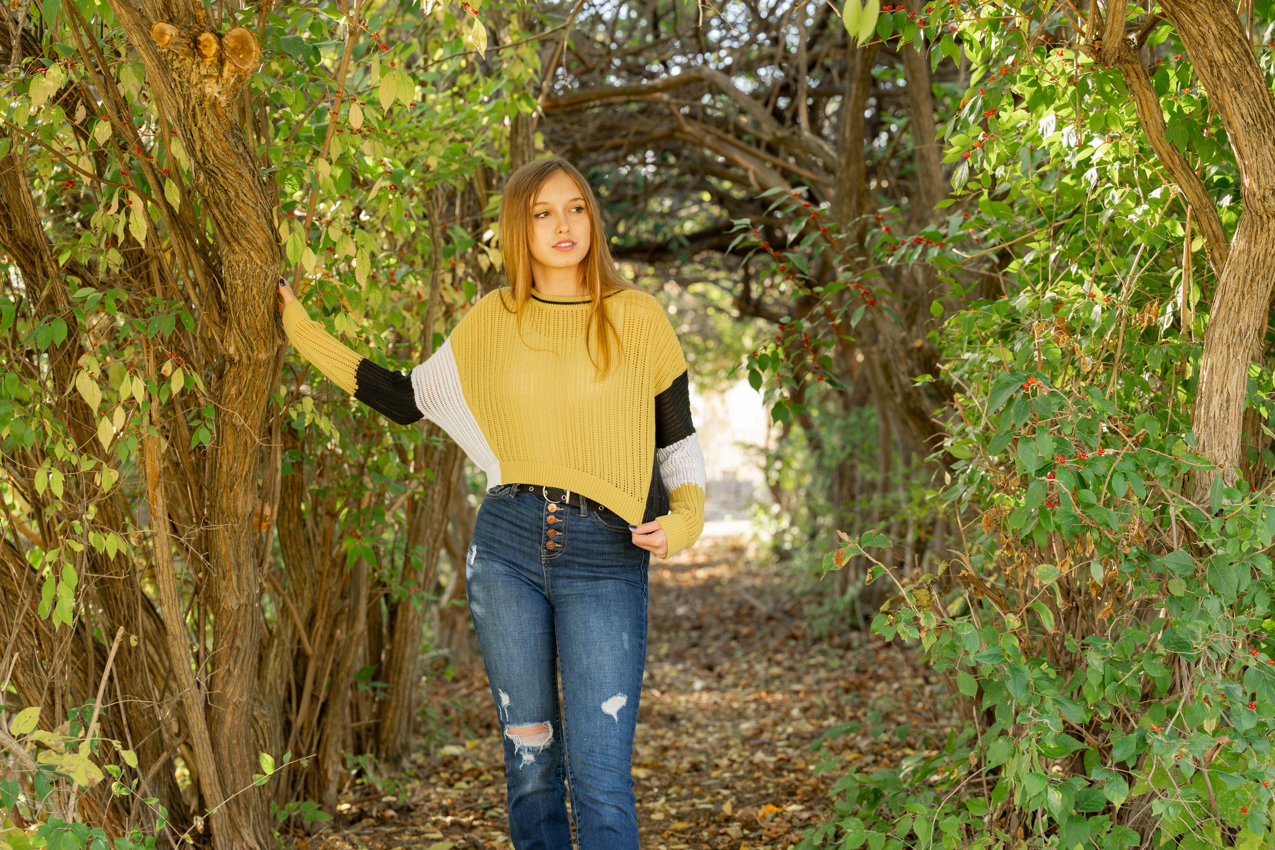 Person in yellow sweater and ripped jeans standing under natural archway of vines.
