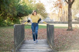 Figure balancing on wooden bridge while wearing yellow sweater and jeans in autumn park setting.