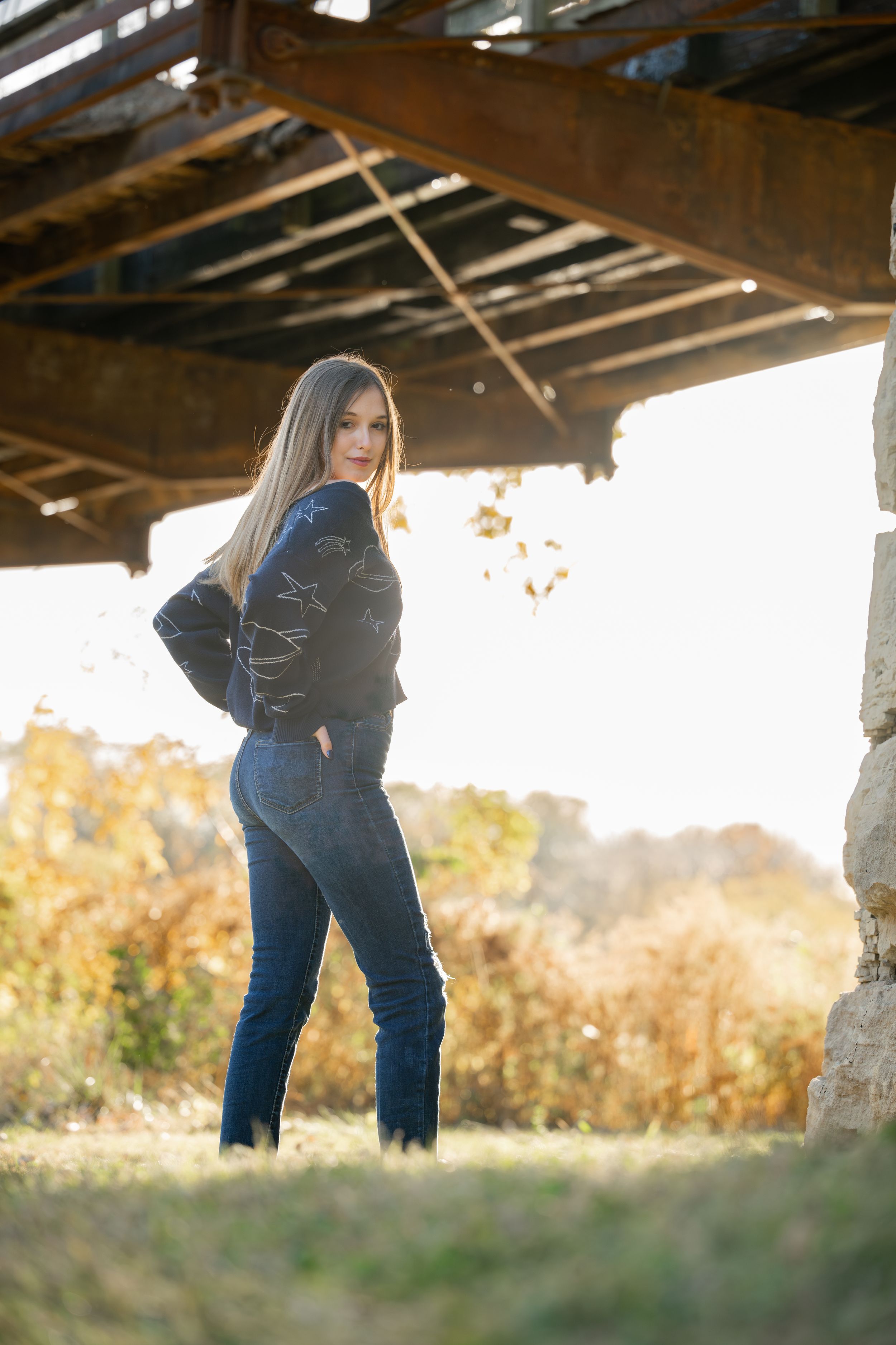 Person posing under rustic wooden structure in blue sweater and jeans at golden hour.
