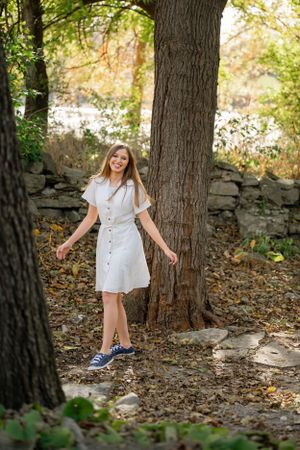 Figure in a white summer dress and sneakers standing by a large tree trunk and stone wall.