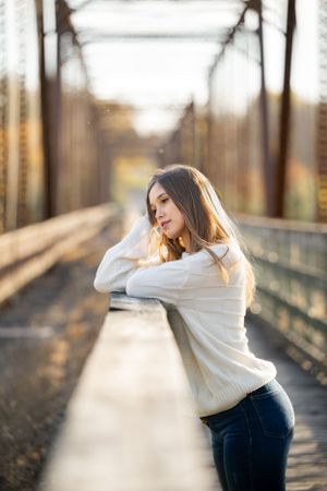 Person in white sweater leaning on metal bridge railing during sunset.