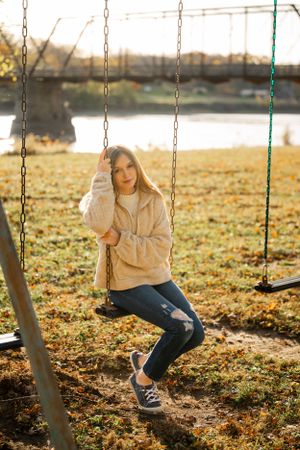 Person in beige jacket sitting on swing set with autumn leaves on ground.