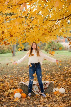 Person in white sweater standing among fall foliage and pumpkins under golden tree.