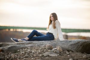 Person in white sweater sitting on rocky shoreline at sunset.