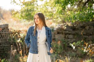 Person wearing denim jacket and white dress poses near rustic stone wall in natural setting.