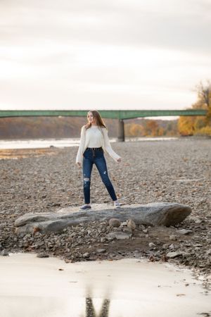 Person standing on large rock by river with bridge in background wearing white sweater.