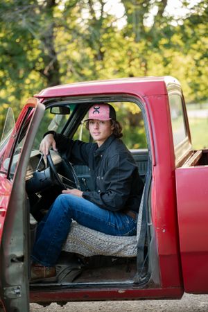 Person sitting in driver's seat of vintage red pickup truck wearing baseball cap and denim.
