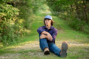 A casual portrait of someone sitting on a forest path wearing denim and baseball cap.