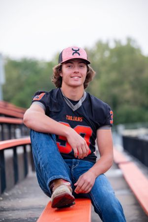Person wearing sports jersey and baseball cap sitting on bleachers.