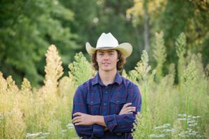 A young person in western wear and cowboy hat stands in a meadow with crossed arms.