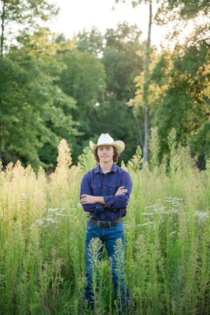 Person in cowboy hat and plaid shirt standing in tall grass field at sunset.