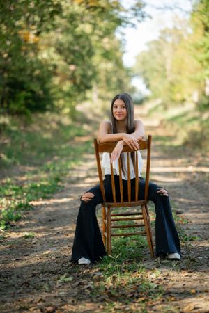 A person sits backwards on a wooden chair on a tree-lined path in autumn.