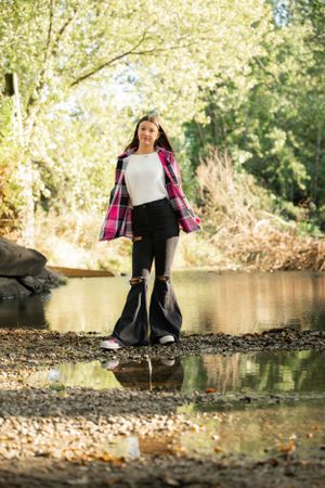 Person in plaid shirt stands at water's edge with reflection in tranquil pond during autumn.