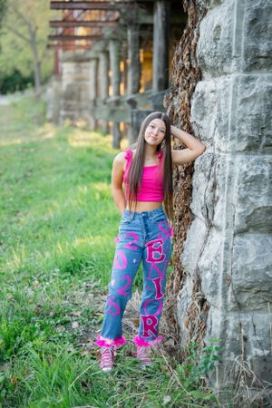 Person in bright pink top poses against stone wall with decorative jeans.