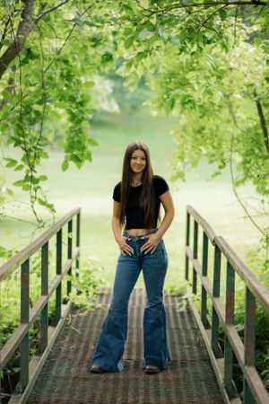 A student poses on a wooden bridge surrounded by lush green foliage near a lake.