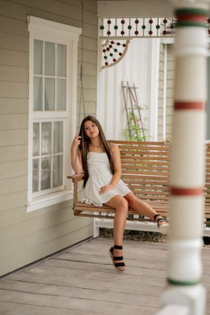 A person in a white dress relaxes on a porch swing by a vintage farmhouse.