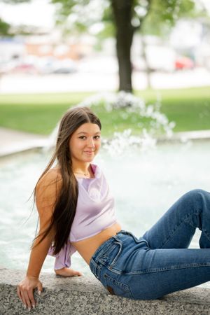 A person in jeans and crop top poses by a fountain on a summer day.