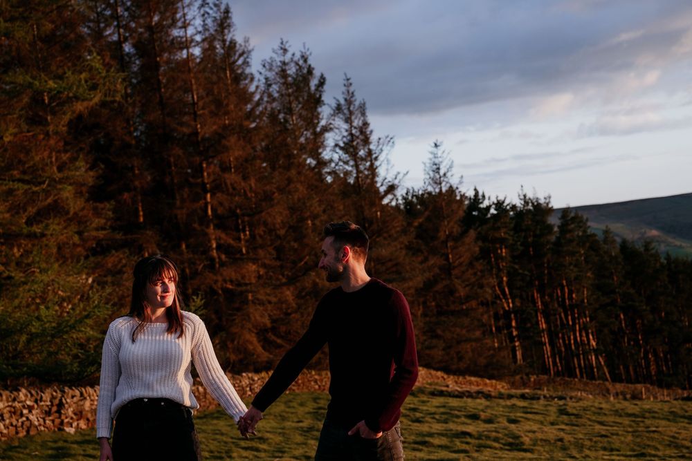 Couple holding hands in a sunlit forest clearing, sharing a romantic moment with mountains in the background.