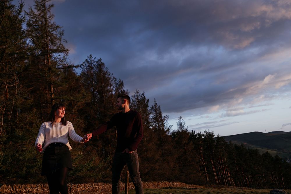 Couple holding hands walking at dusk with dramatic purple clouds and forest silhouette in the background.