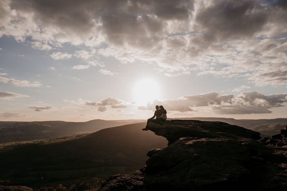 Two people sit together on a rocky hilltop at sunset, overlooking a vast scenic landscape under a dramatic cloudy sky.