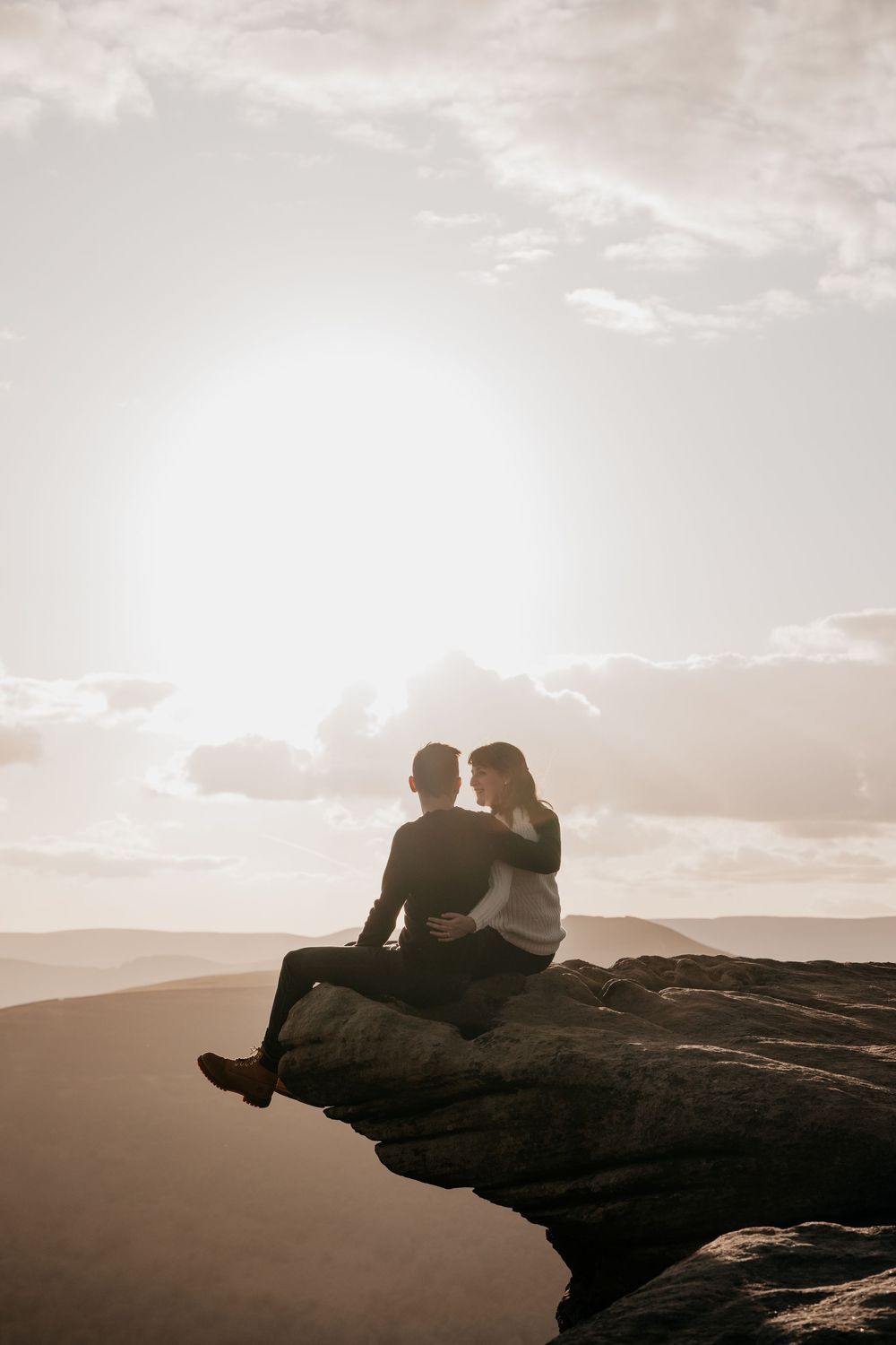 Couple sitting together on a rocky cliff edge at sunset, embracing romantically with mountain views.