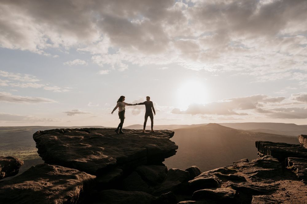 Couple holding hands on rocky mountain peak at sunset with dramatic cloudy sky and valley views.