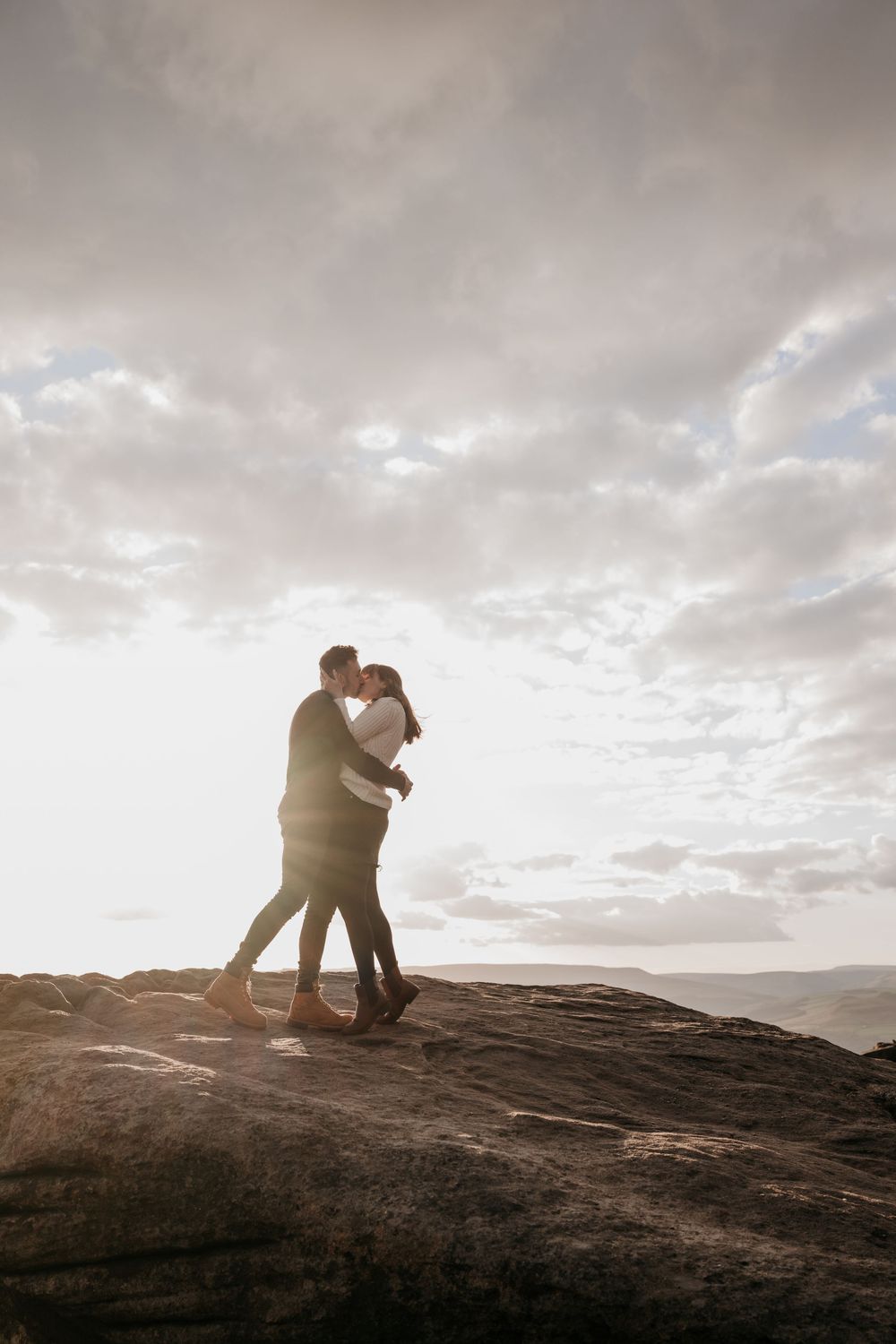 Couple embracing and kissing on a rocky mountain summit at sunset with dramatic cloudy sky backdrop.