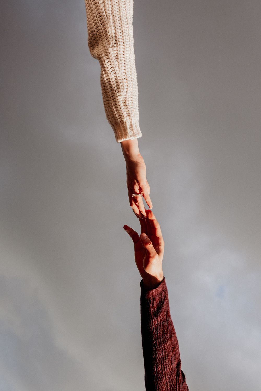 Two hands reaching toward each other against a gray sky, one in a cream knit sweater and one in a burgundy sleeve.