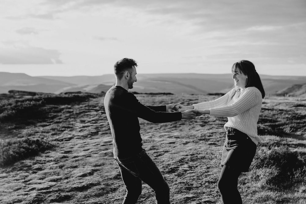 Black and white photo of a couple holding hands on a hilltop with scenic moorland landscape behind them.
