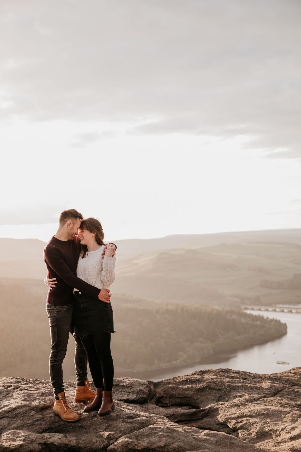 Romantic couple embracing on a rocky cliff edge at sunset with a river valley stretching behind them.