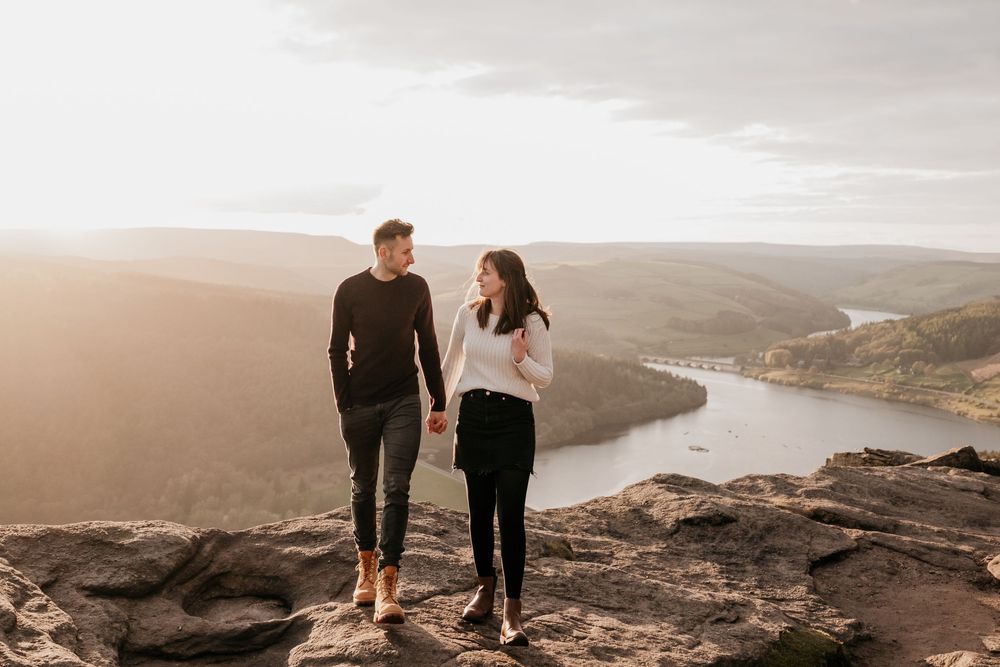Happy couple holding hands walking on a rocky summit with a stunning river and valley view at golden hour.