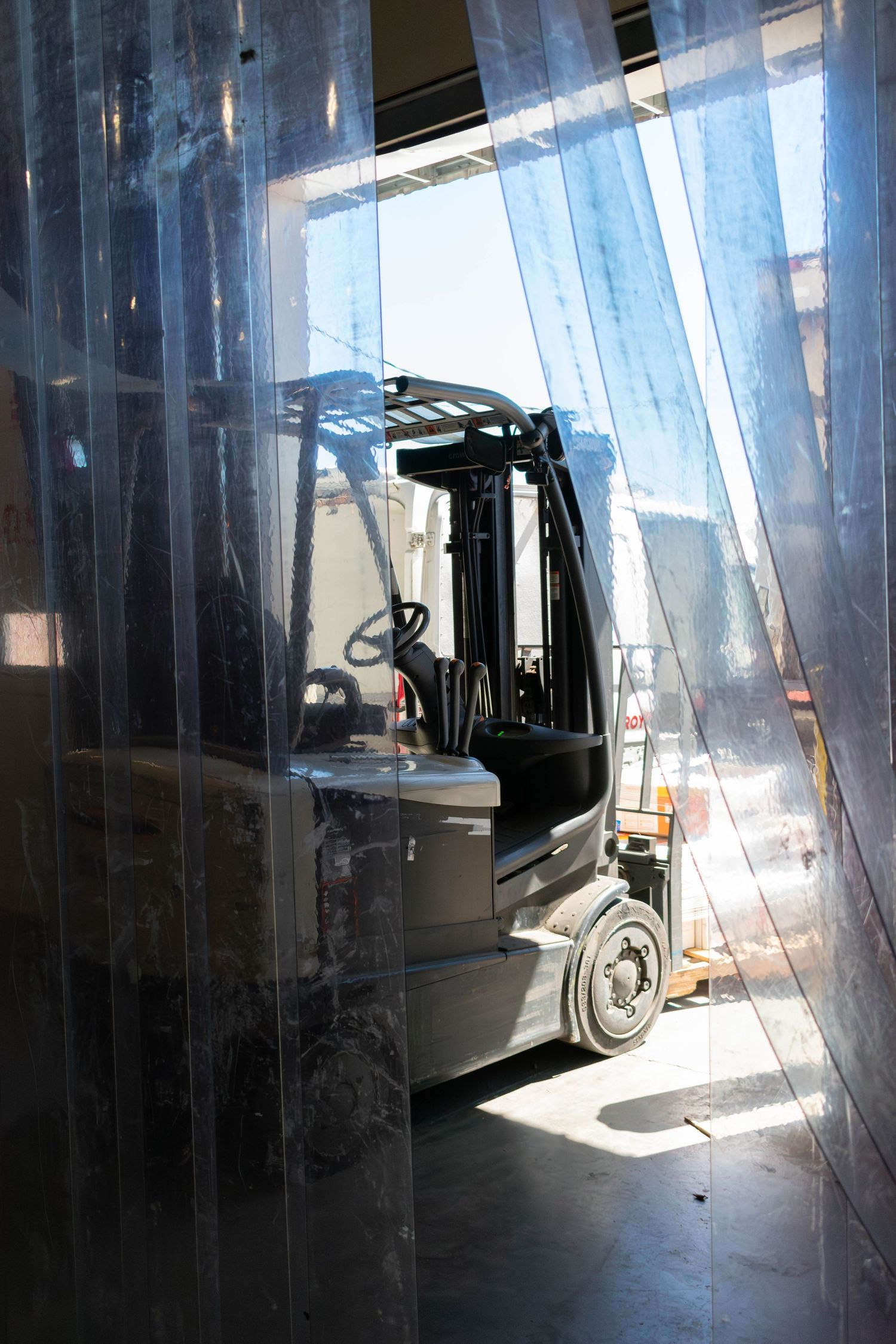 Forklift visible through translucent plastic curtains with sunlight creating dramatic shadows.