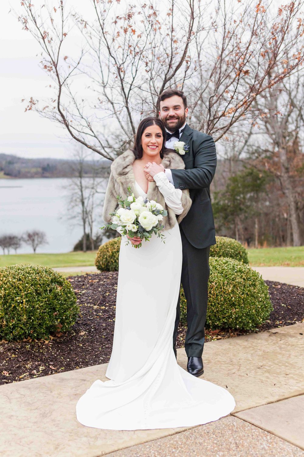 A winter wedding couple poses outdoors by a lake with bare trees and shrubs in the background.