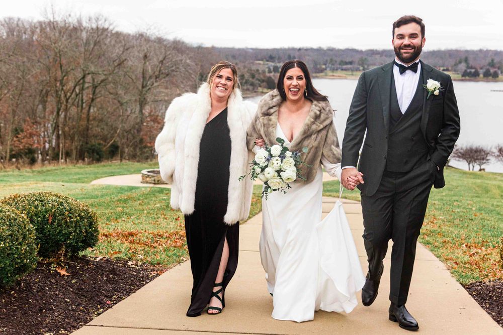 Three people walking outdoors at a winter wedding, with bride in white gown holding bouquet and wearing fur shawl.
