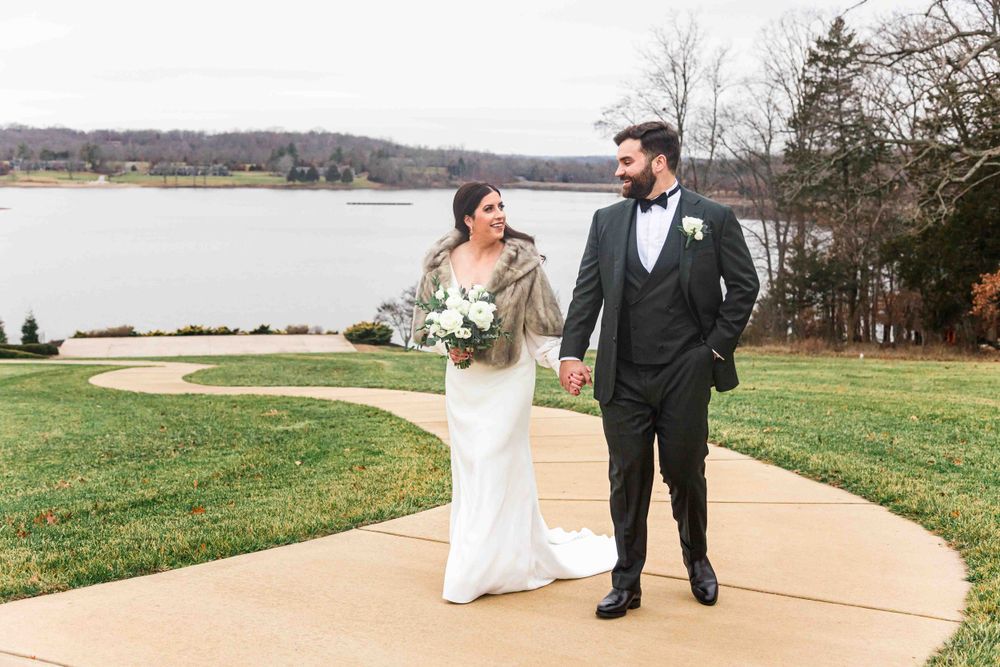 A couple walks together outdoors along a lakeside path on a cloudy day, with bride in white gown and groom in dark suit.