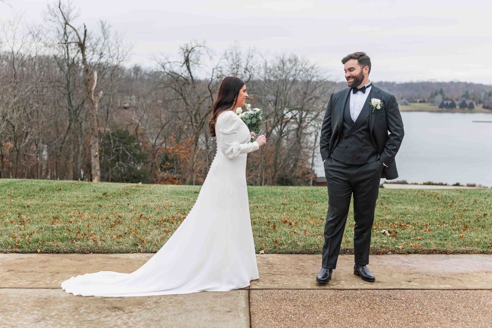 A romantic outdoor wedding scene with a couple in formal attire standing by a serene lake view on an autumn day.