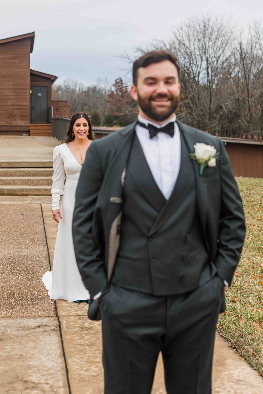 Wedding first look moment with bride in long sleeve dress and groom in black tuxedo at an outdoor venue.