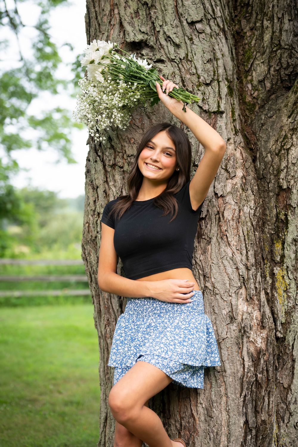 A person in a black top and floral skirt poses against a large tree trunk while holding white wildflowers overhead.