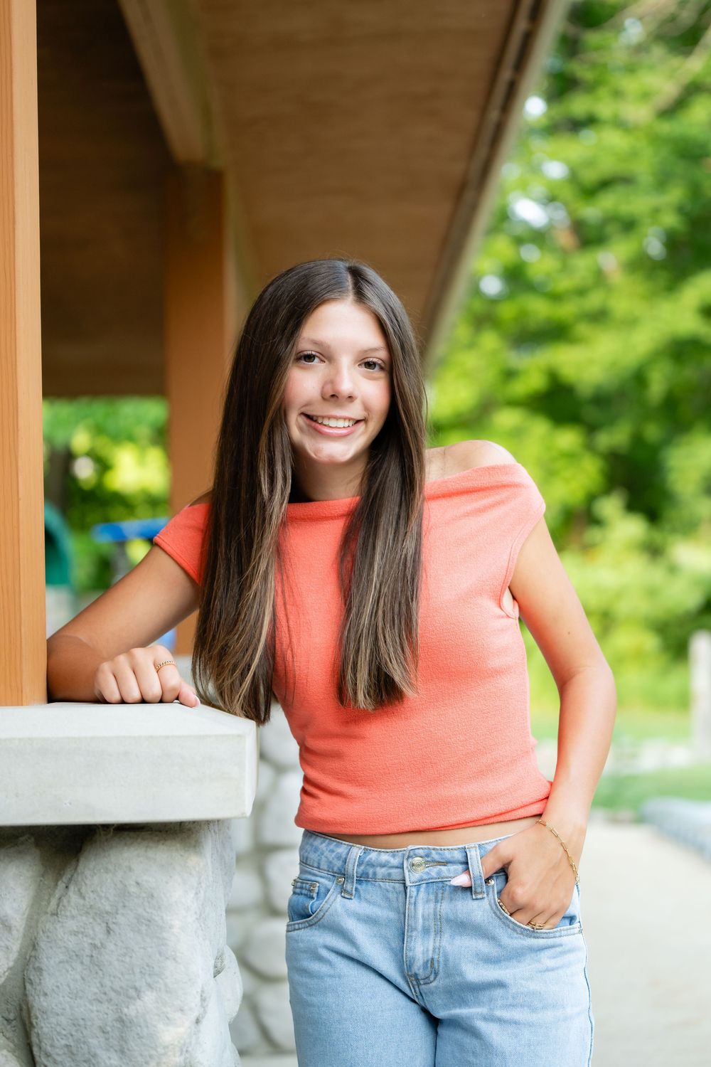 Smiling teenage girl with long brown hair wearing coral top and jeans, leaning against stone pillar on outdoor porch.